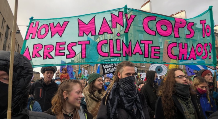 A crowd of protesters under a banner saying "How many cops to arrest climate chaos?"