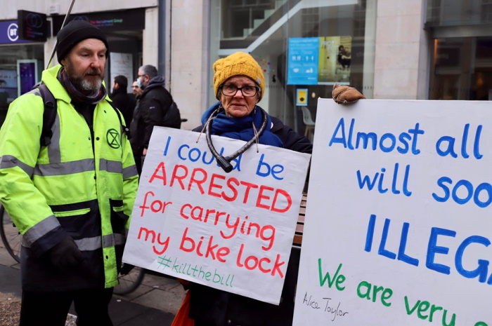 A woman with a bike lock around her neck wears a placard which says
I could be arrested for carrying my bike lock
#killthebill