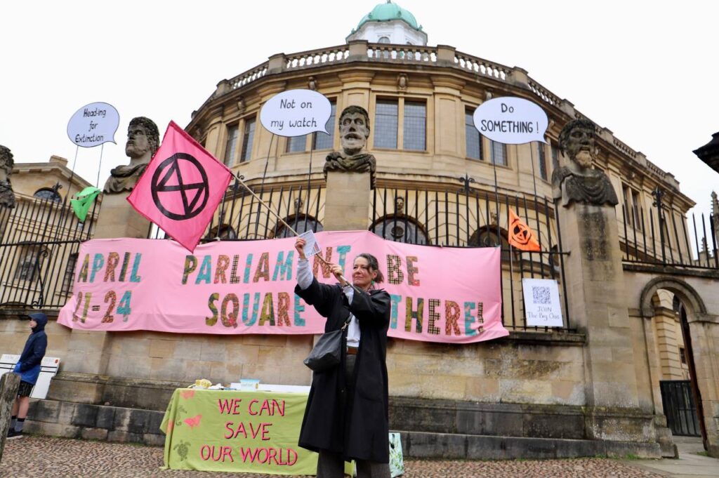 A woman waves a pink XR flag in front of the Sheldonian in Oxford. The famous heads have been given speech bubbles reading
"Heading for Extinction"
"Not on my watch"
"Do SOMETHING!"
A banner is attached to the railings reading:
"April 21-24 Parliament Square Be There!"